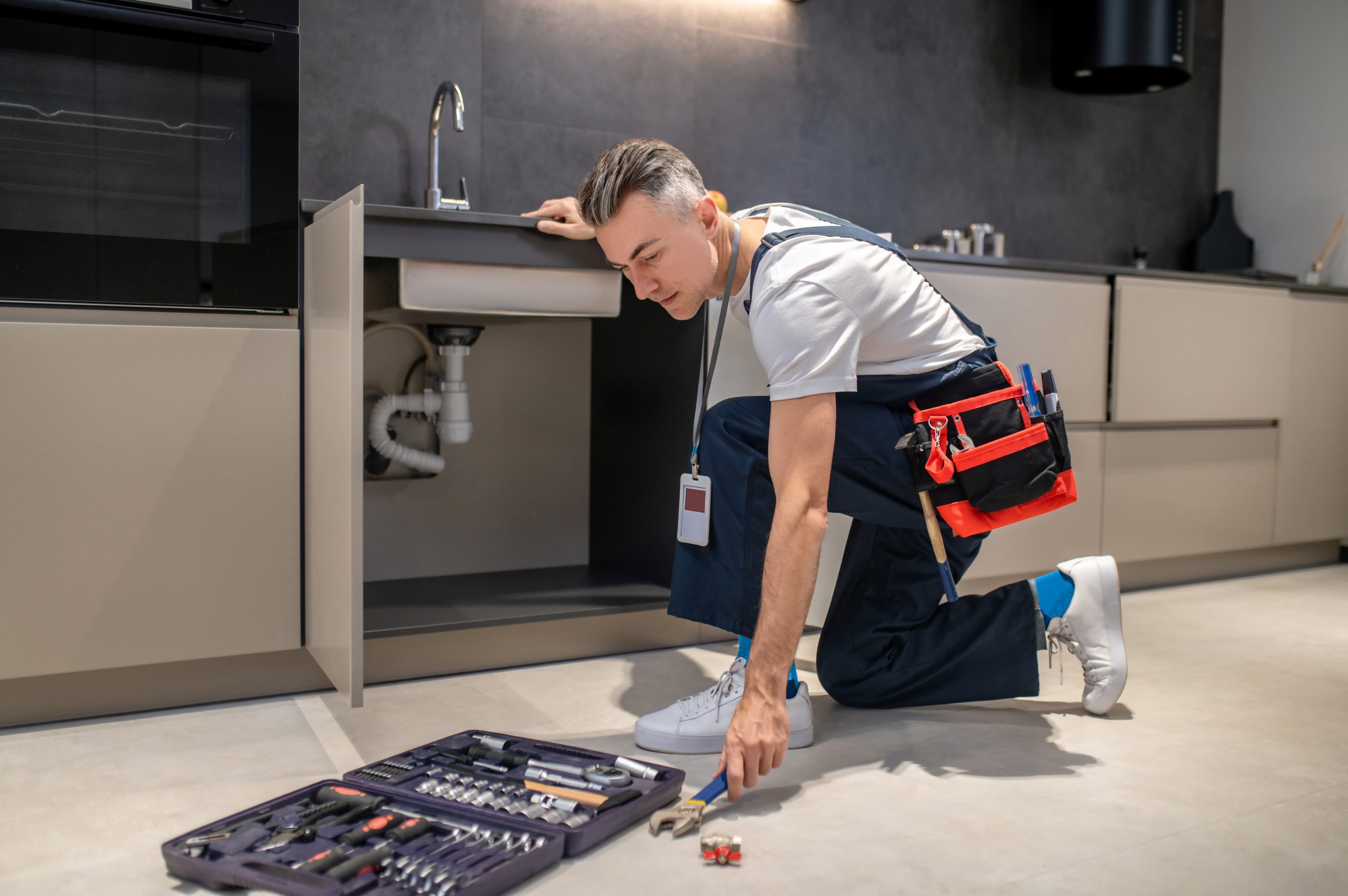 man crouched near sink stretching out hand to wrench