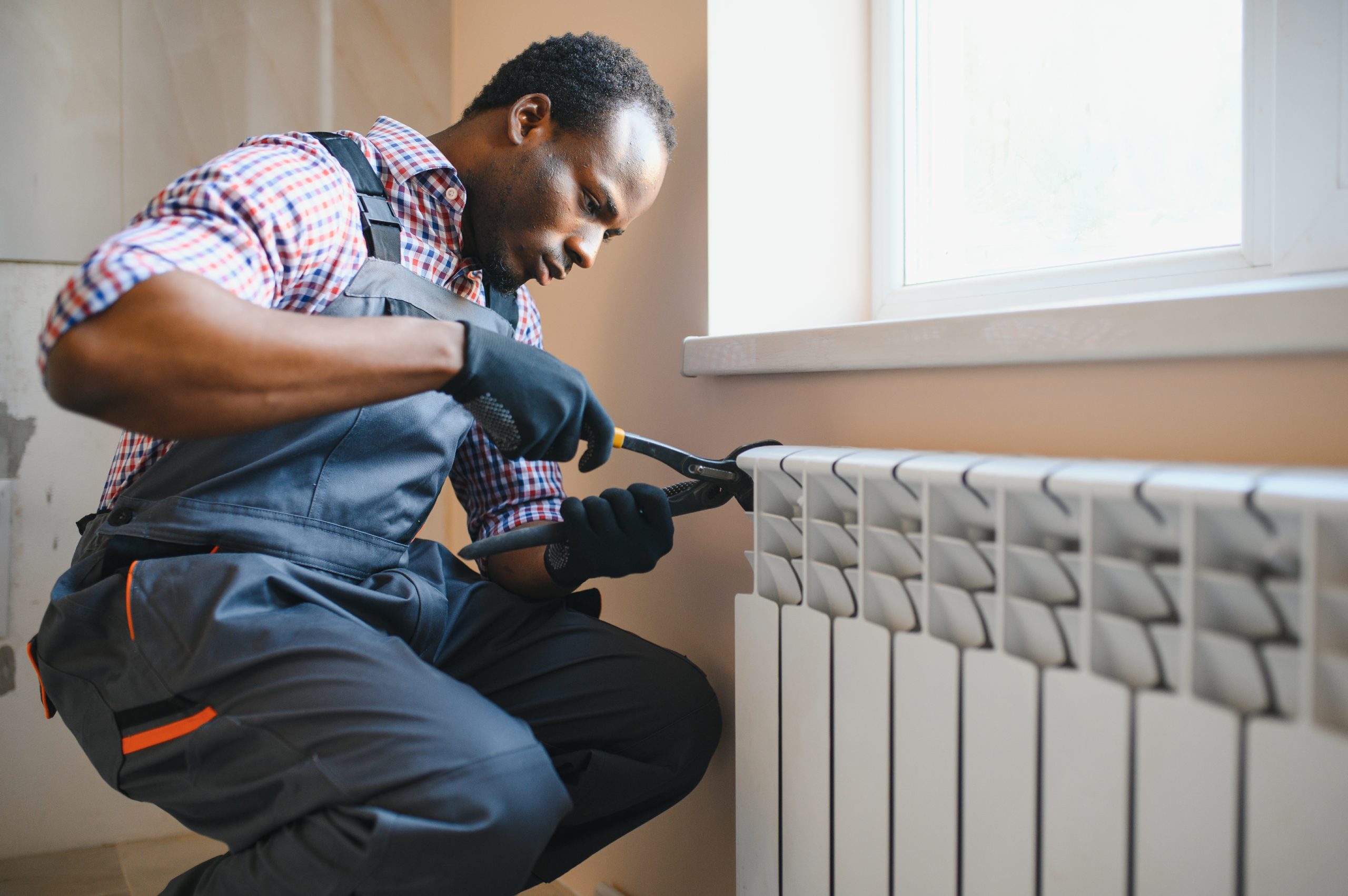 man in workwear overalls using tools while installing or repairing heating radiator in room