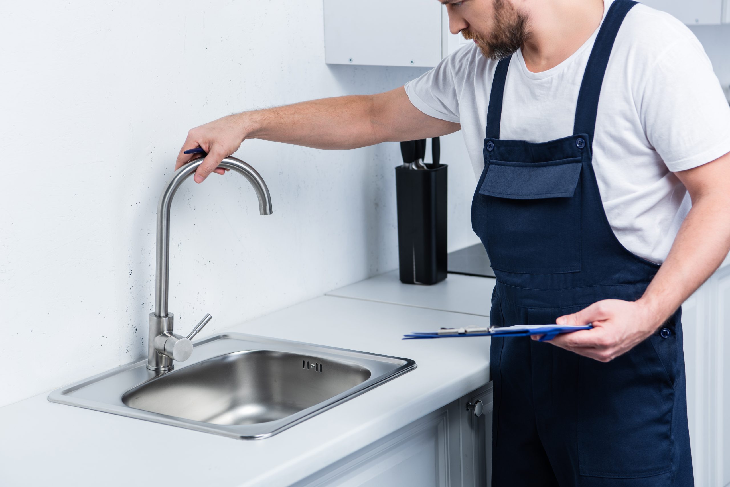 partial view of repairman in working overall holding clipboard and checking sink in kitchen